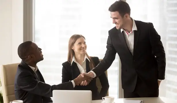 cheerful businessmen handshaking while female colleague watches smiling