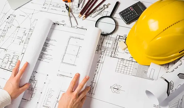 hands unrolling a construction plan blueprint with a yellow hardhat, calculator, protractor, and magnifying glass in the background