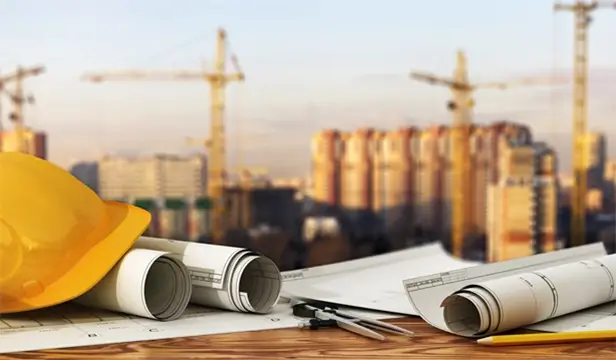 yellow hardhat and construction plans on a table with tall buildings and cranes in the background